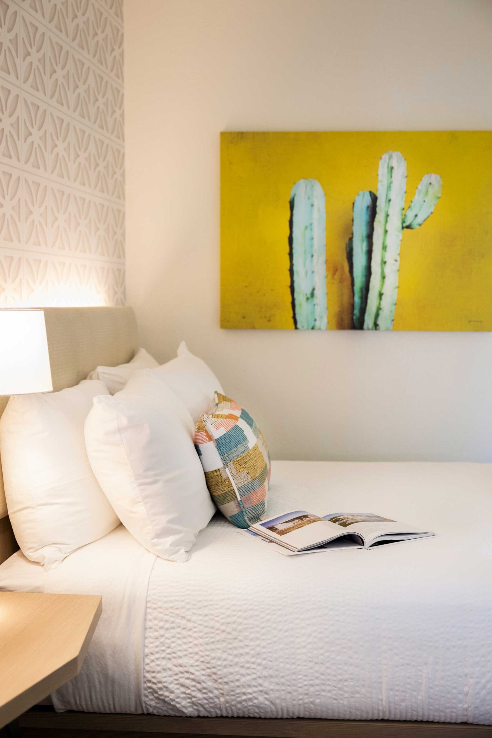 White bedding with fluffy pillows sits on a bed next to a lamp; a bright yellow cactus painting hangs on the wall behind.