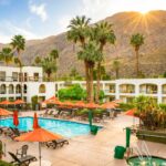 Pool area at a sunny resort with orange umbrellas, lounge chairs, palm trees, and white buildings against a mountainous backdrop.