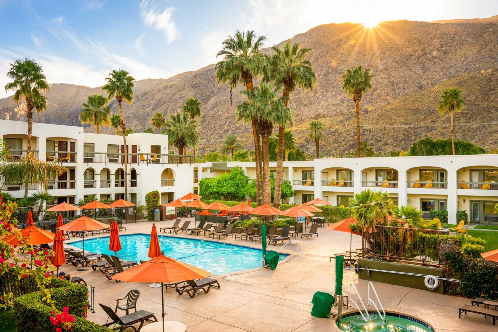 Pool area at a sunny resort with orange umbrellas, lounge chairs, palm trees, and white buildings against a mountainous backdrop.