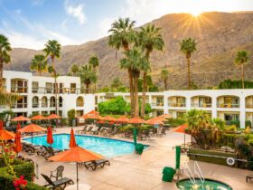 Pool area at a sunny resort with orange umbrellas, lounge chairs, palm trees, and white buildings against a mountainous backdrop.