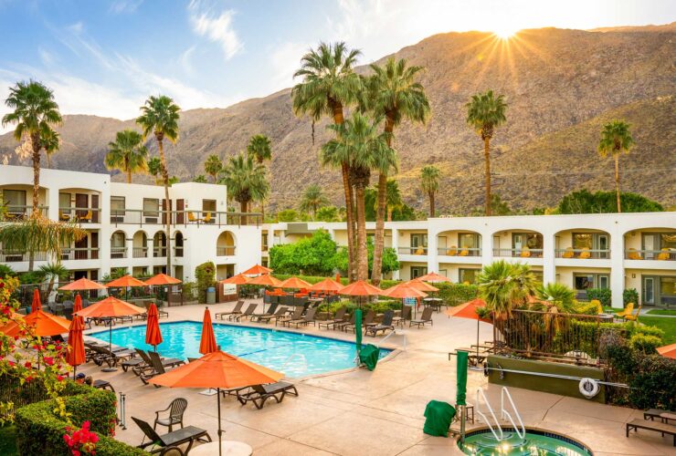Pool area at a sunny resort with orange umbrellas, lounge chairs, palm trees, and white buildings against a mountainous backdrop.