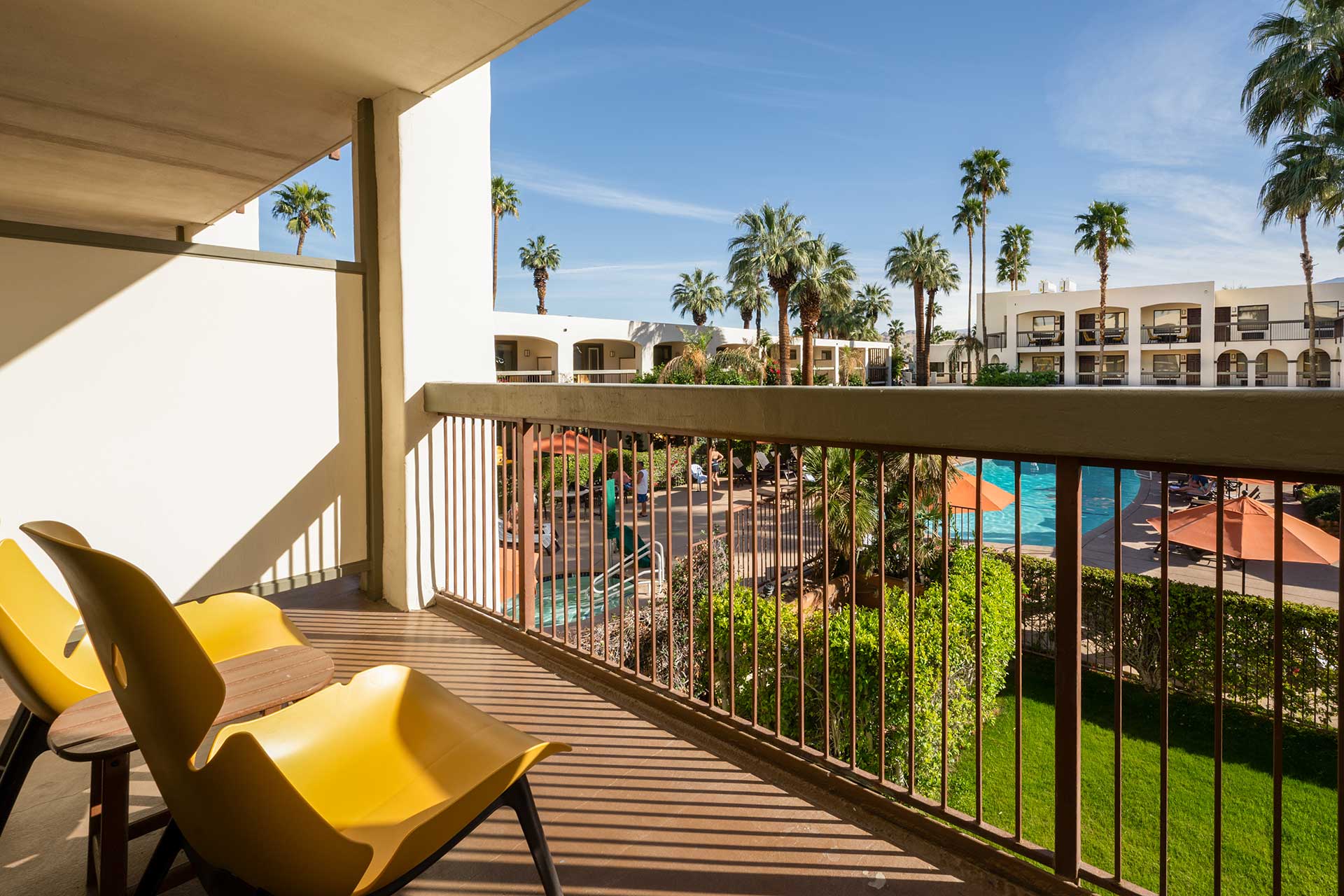 Balcony with yellow lounge chairs overlooking a pool and palm trees at a sunny resort.
