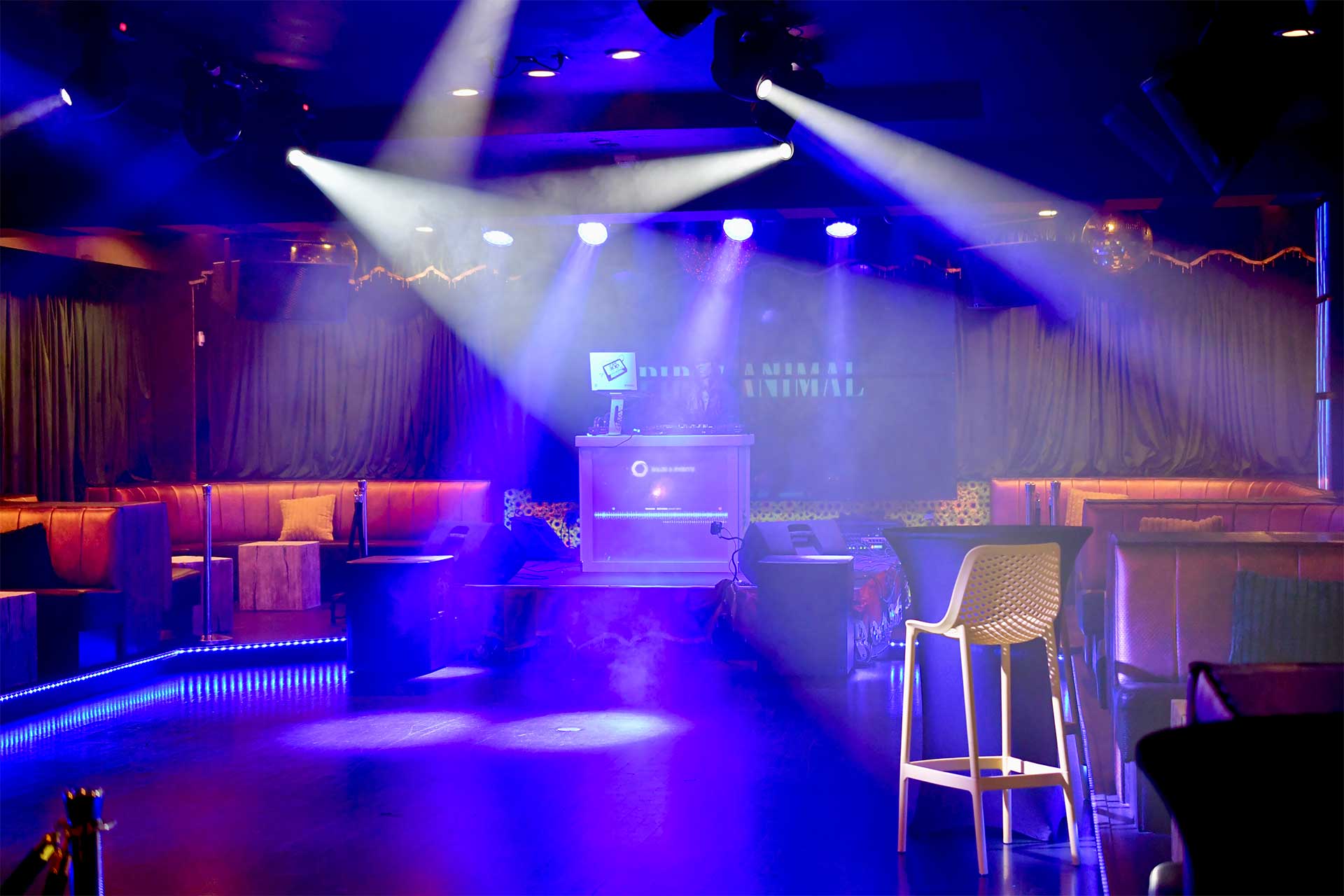 Nightclub stage with blue and purple lighting, a DJ booth at center with a laptop, surrounded by lounge seating and hazy atmosphere.