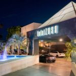 Night exterior of Hotel Zoso with a pyramid roof, illuminated sign, palm trees, and a lit fountain in the foreground.