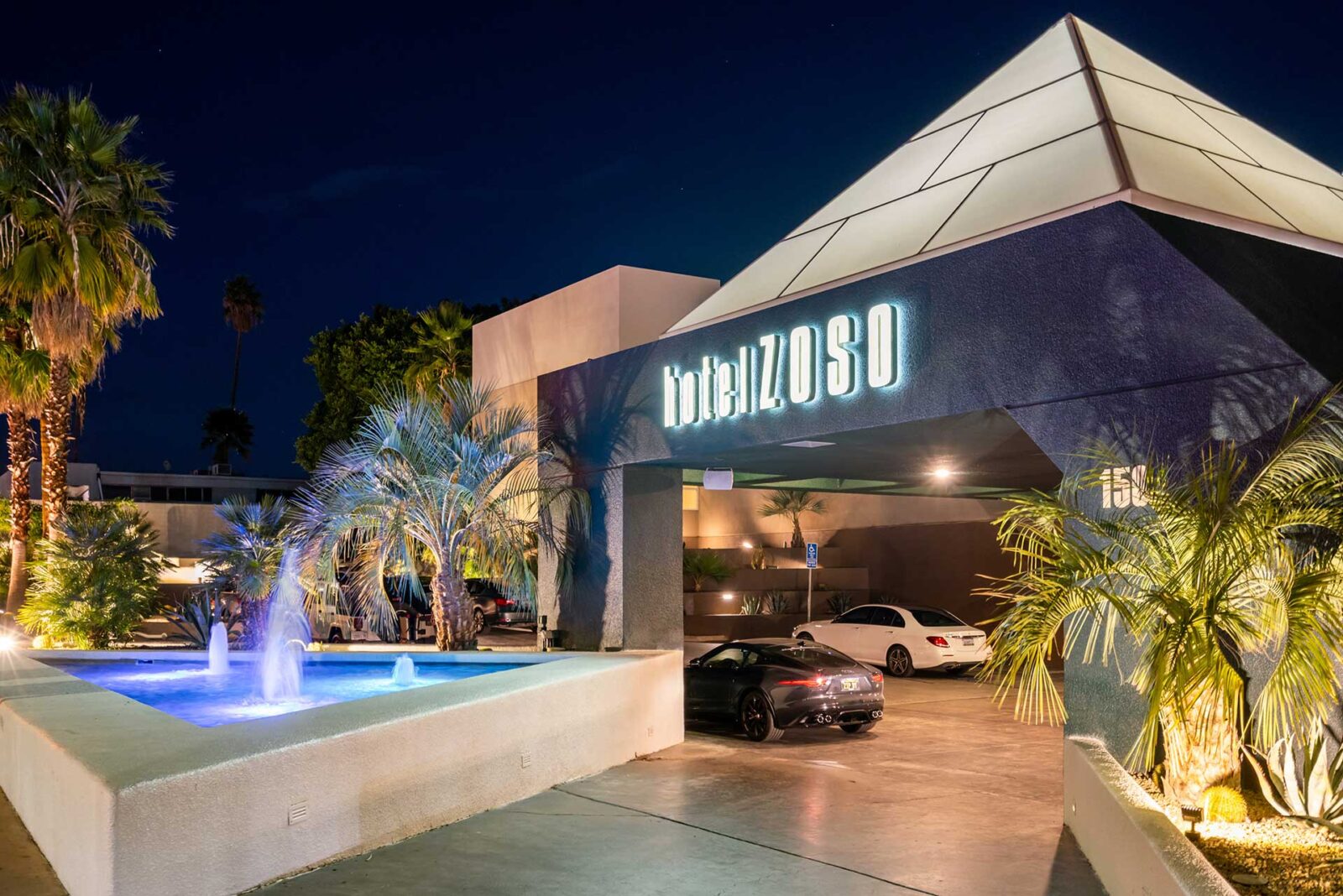 Night exterior of Hotel Zoso with a pyramid roof, illuminated sign, palm trees, and a lit fountain in the foreground.