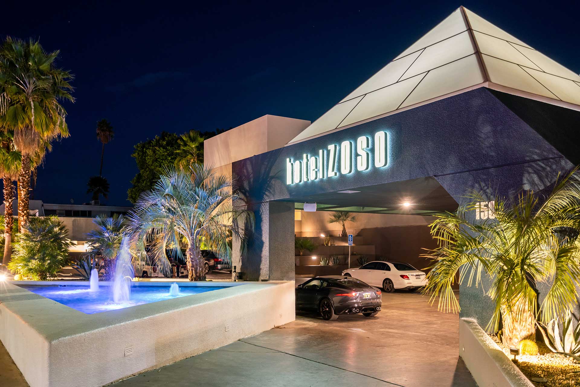 Night exterior of Hotel Zoso with a pyramid roof, illuminated sign, palm trees, and a lit fountain in the foreground.