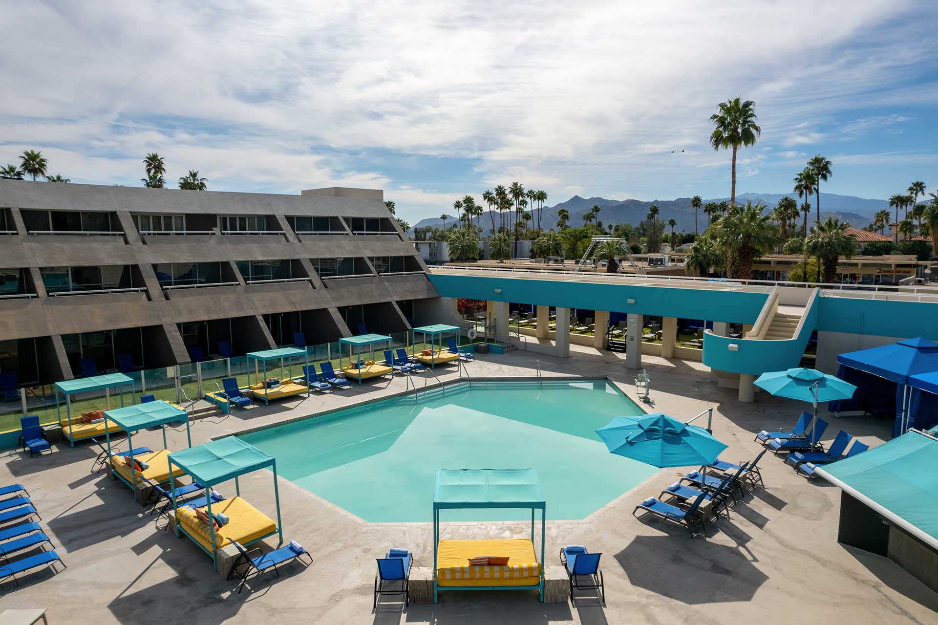Poolside at a midcentury modern hotel: turquoise pool, blue and yellow lounge chairs under teal canopies, and palm trees with distant mountains.
