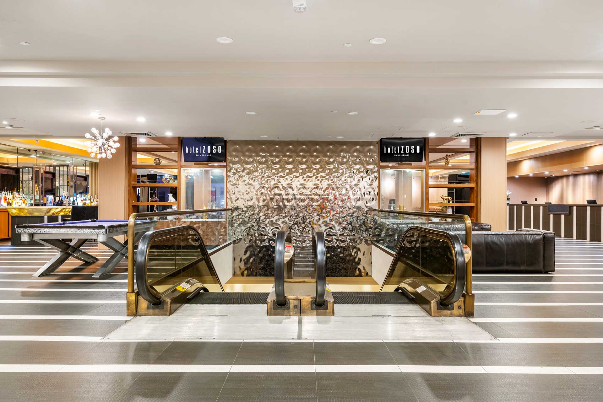 Hotel lobby with two glass-paneled escalators flanking a wavy metallic screen; bar area to the left and reception to the right.