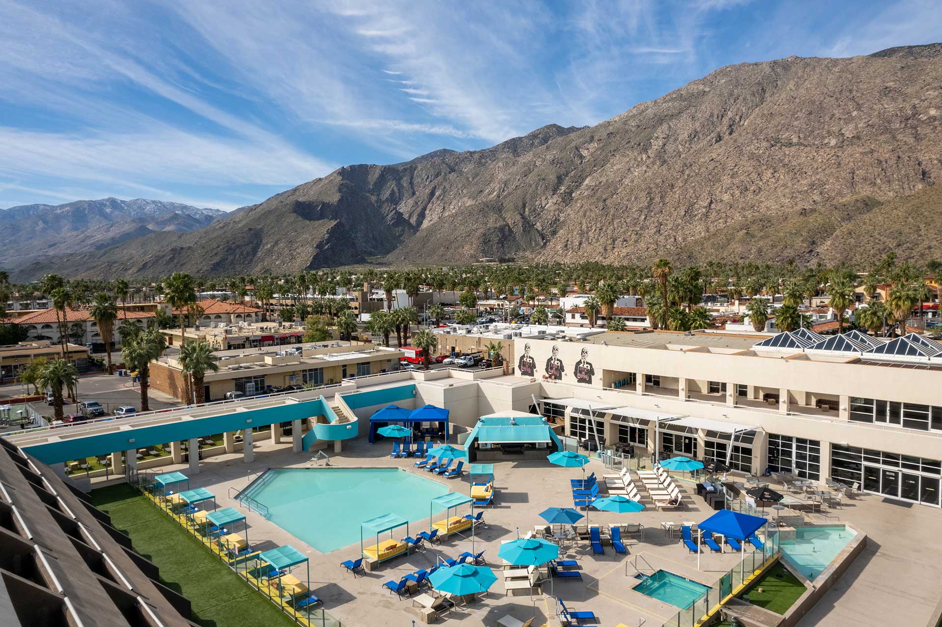 Resort pool deck with blue umbrellas and lounge chairs, palm trees, and a distant rocky mountain range.