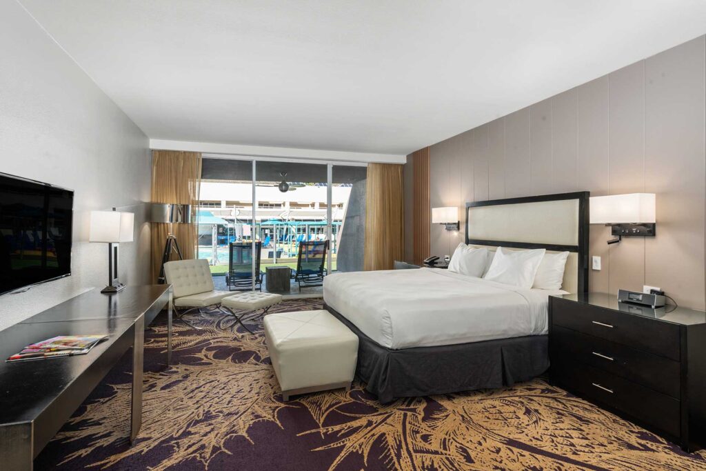 Hotel room with a king bed, seating area, and sliding glass doors opening to a pool-view balcony.