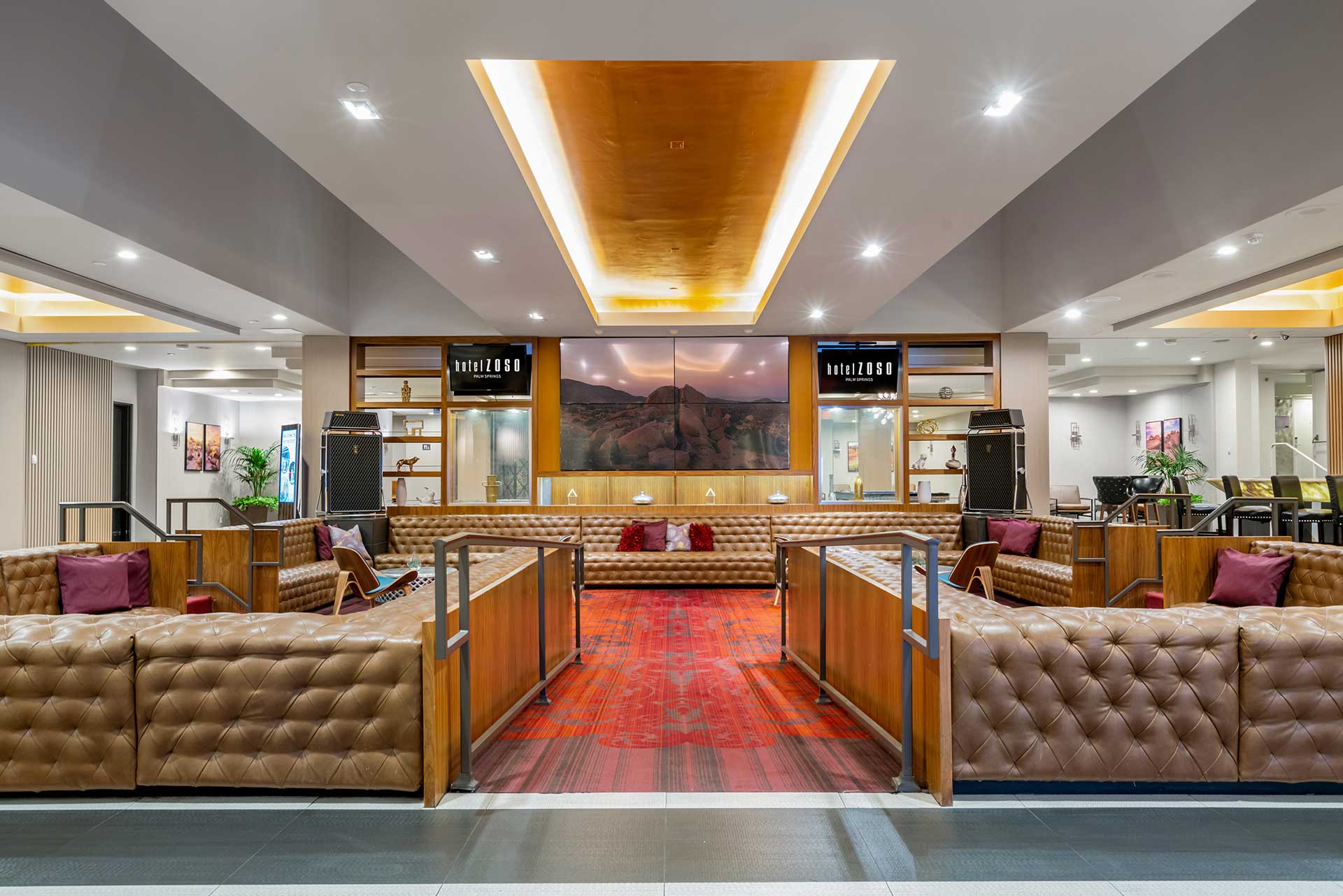 Hotel lobby with wide, brown tufted sofas, red carpet, and a central reception area with screens above a mural.